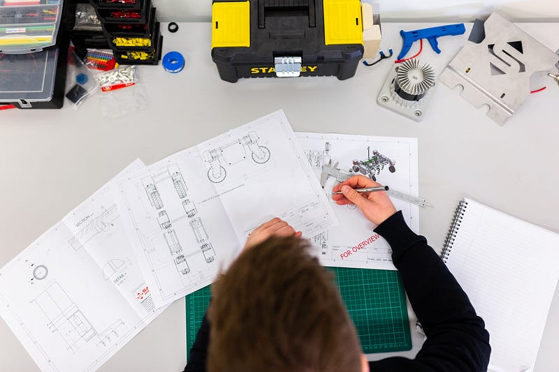 Top-down view of an engineer examining technical blueprints and schematics for a robotics project at a workbench filled with tools, parts organizers, and a toolbox.