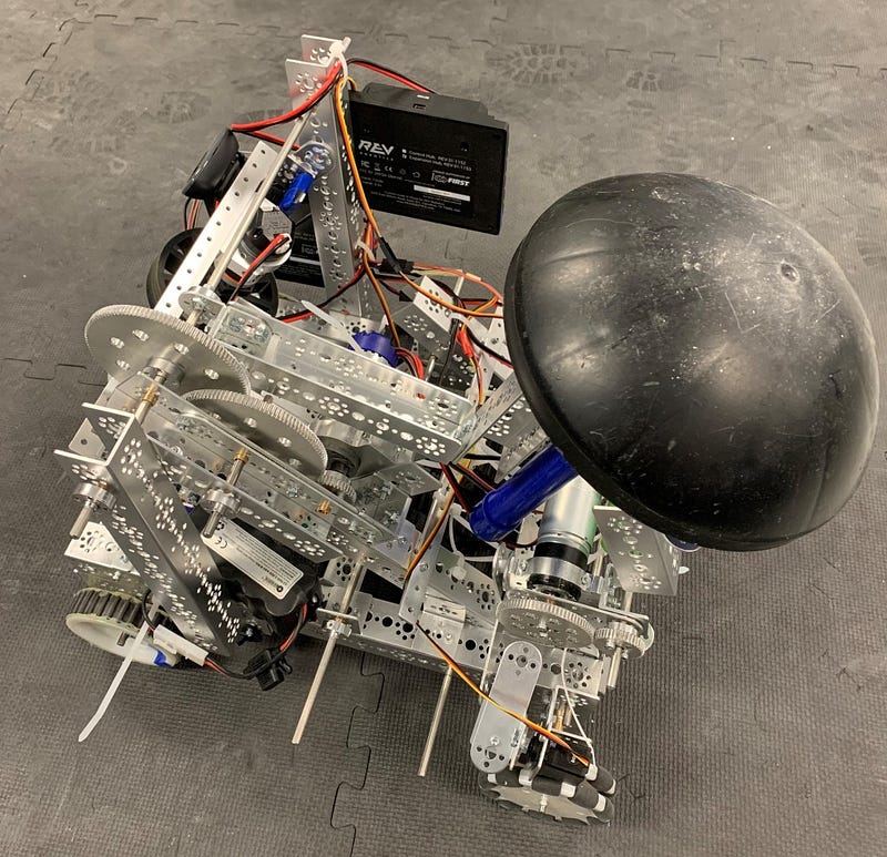 High-angle photograph of a custom-built robotics competition robot built with aluminum channel framing, gears, omni-directional wheels, REV Robotics control modules, exposed wiring, and a large black dome-shaped launcher attachment.