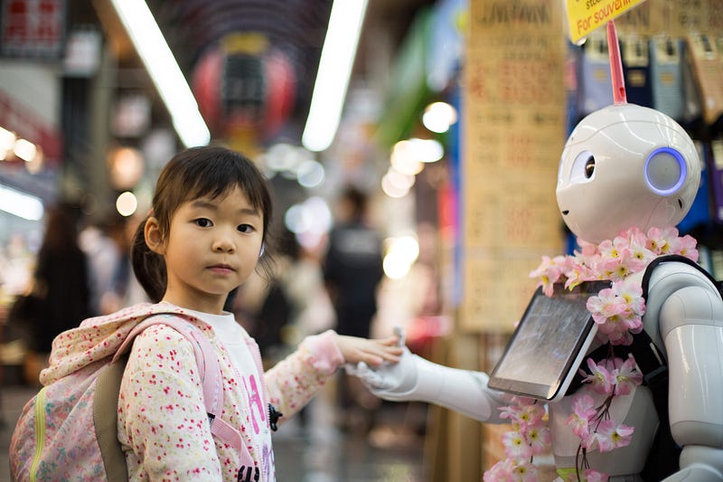 A young girl wearing a patterned sweater and backpack holding hands with a Pepper humanoid robot decorated with pink cherry blossom garlands in a busy indoor marketplace.