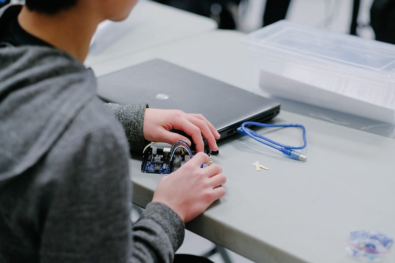 Close-up photograph of a student in a grey hoodie connecting jumper wires to a microcontroller circuit board next to a laptop on a desk.