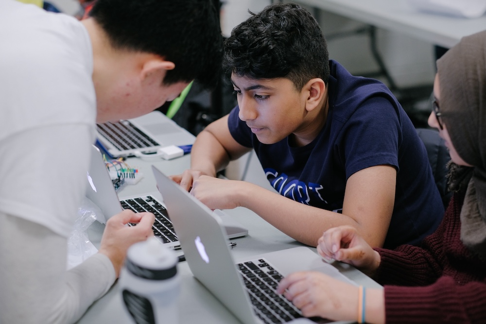 A young male student focused on a laptop screen while an instructor leans in to assist him, alongside a classmate working on her computer at the same desk