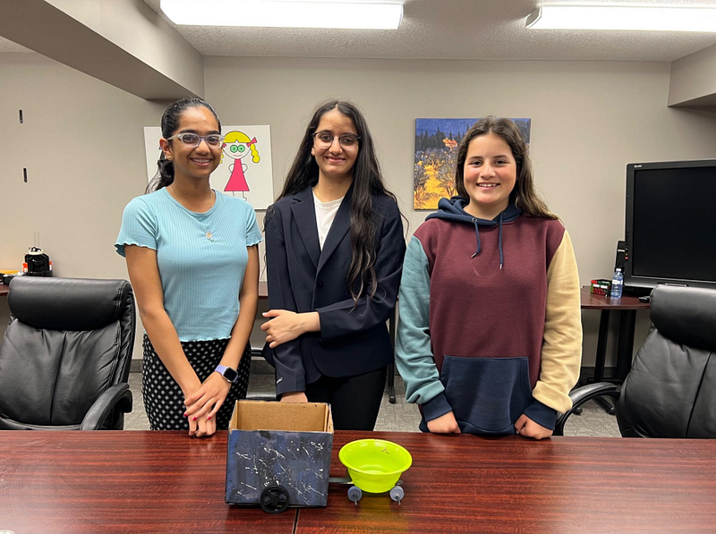 Three smiling young female students standing behind a wooden conference table presenting a handmade prototype made from a painted cardboard box and a green plastic bowl mounted on a wheeled chassis.