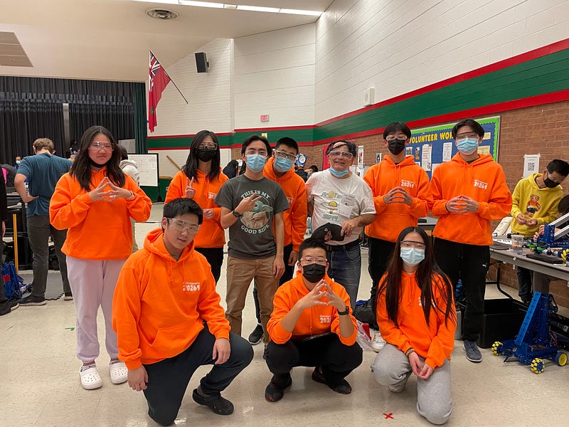 Group portrait of a robotics team wearing matching bright orange hoodies, safety glasses, and face masks posing together in a school gymnasium at a competition event.