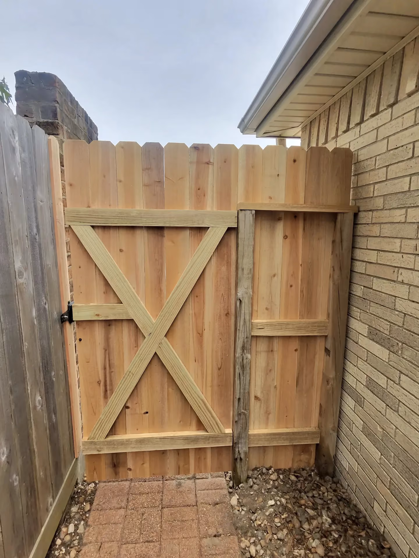 A cedar dog-ear gate with a diagonal X-brace meets a brick house corner on the Texas Gulf Coast.
