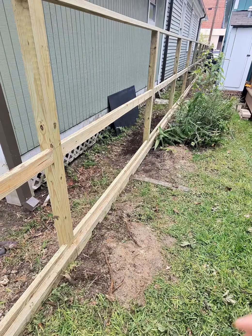 New pressure-treated fence rails run along a side yard beside pale green siding, with fresh lumber staged on the ground on the Texas Gulf Coast.
