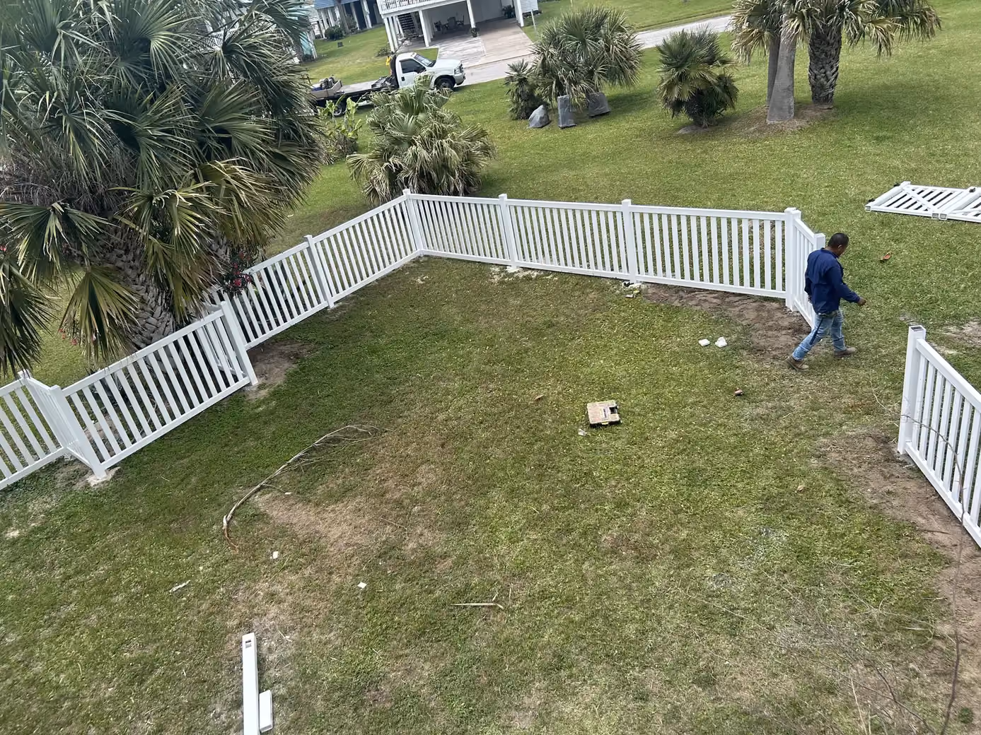 An aerial view shows a white vinyl picket fence enclosure being assembled with gate panels laid nearby in Houston, TX.