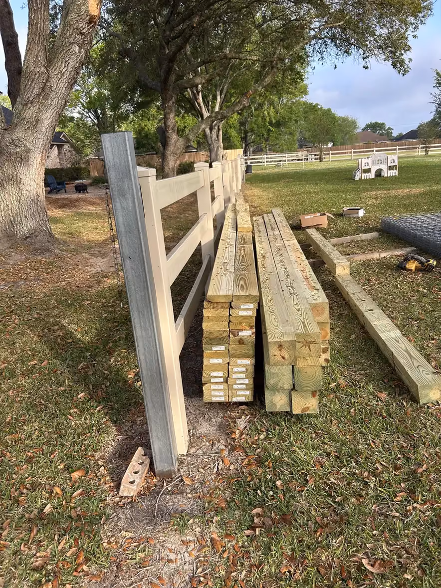 Stacks of treated lumber and posts lean against a finished white and tan rail fence in Houston, TX.