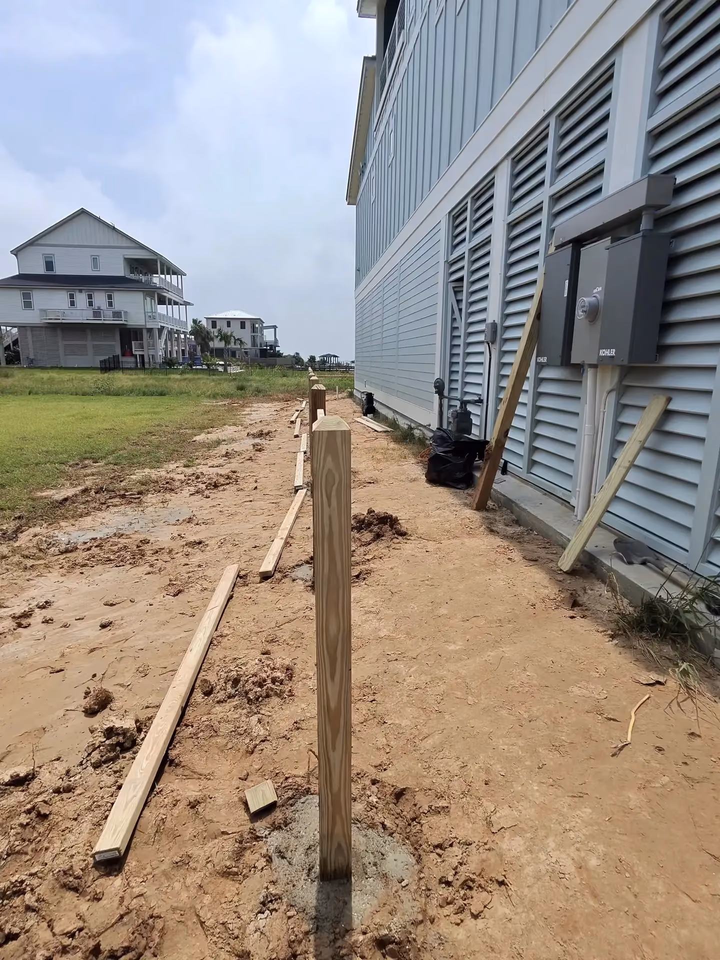 A single pressure-treated fence post freshly set in muddy soil beside an elevated blue coastal home in Houston, TX.