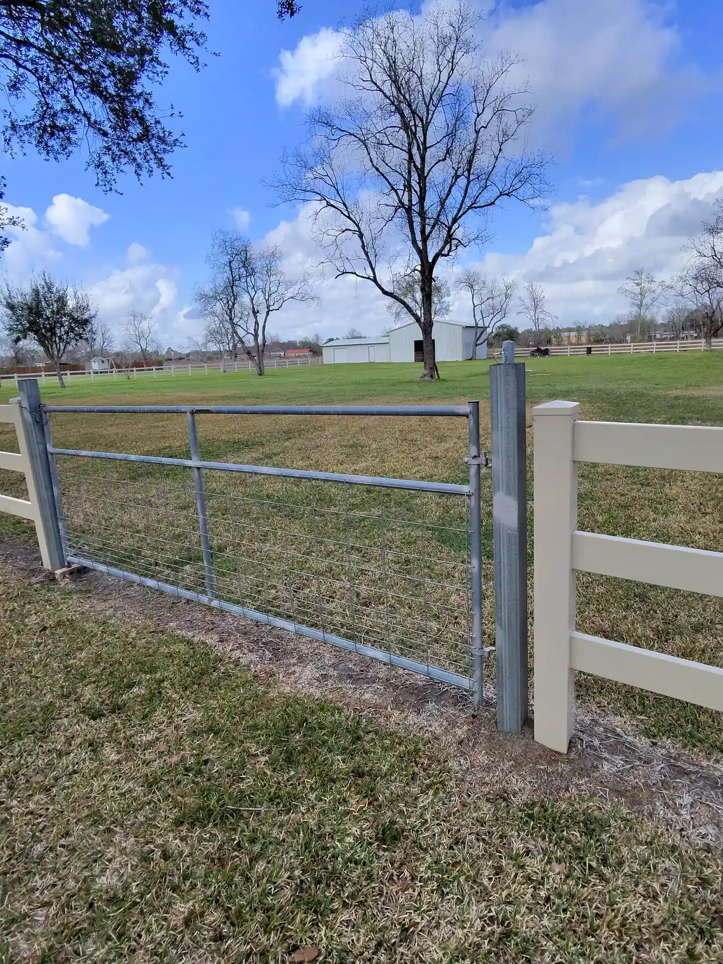 A tan vinyl three-rail fence connecting to a galvanized metal wire gate on a rural property with a barn in Houston, TX.