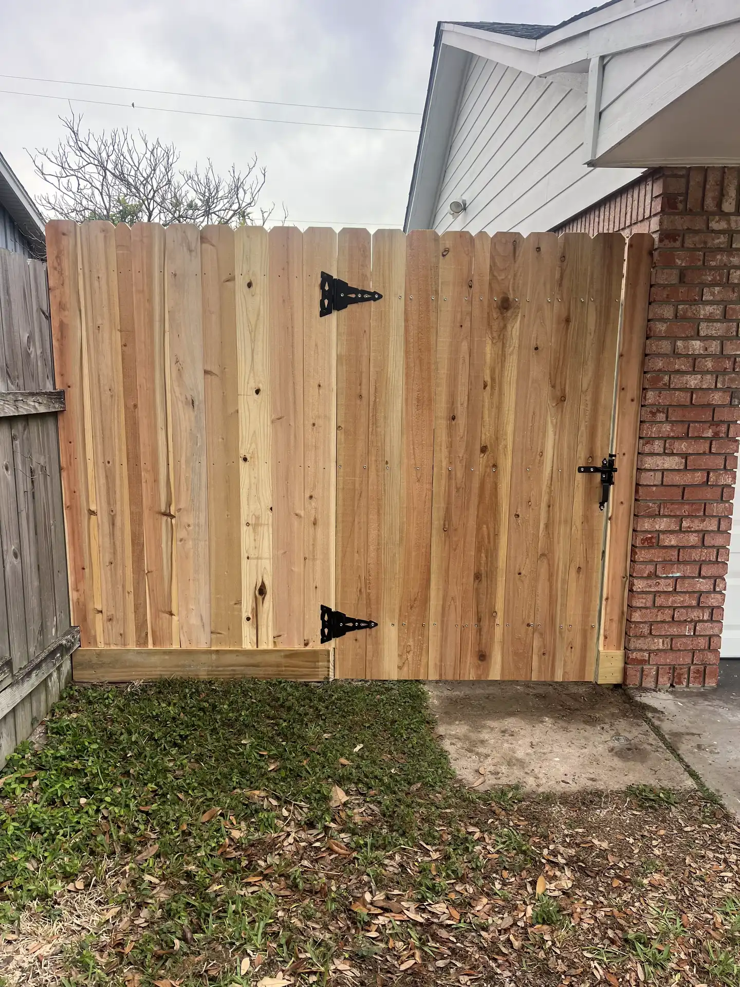 Natural cedar gate with black T-hinges and latch set into a wood fence beside a brick garage in Galveston County, TX.