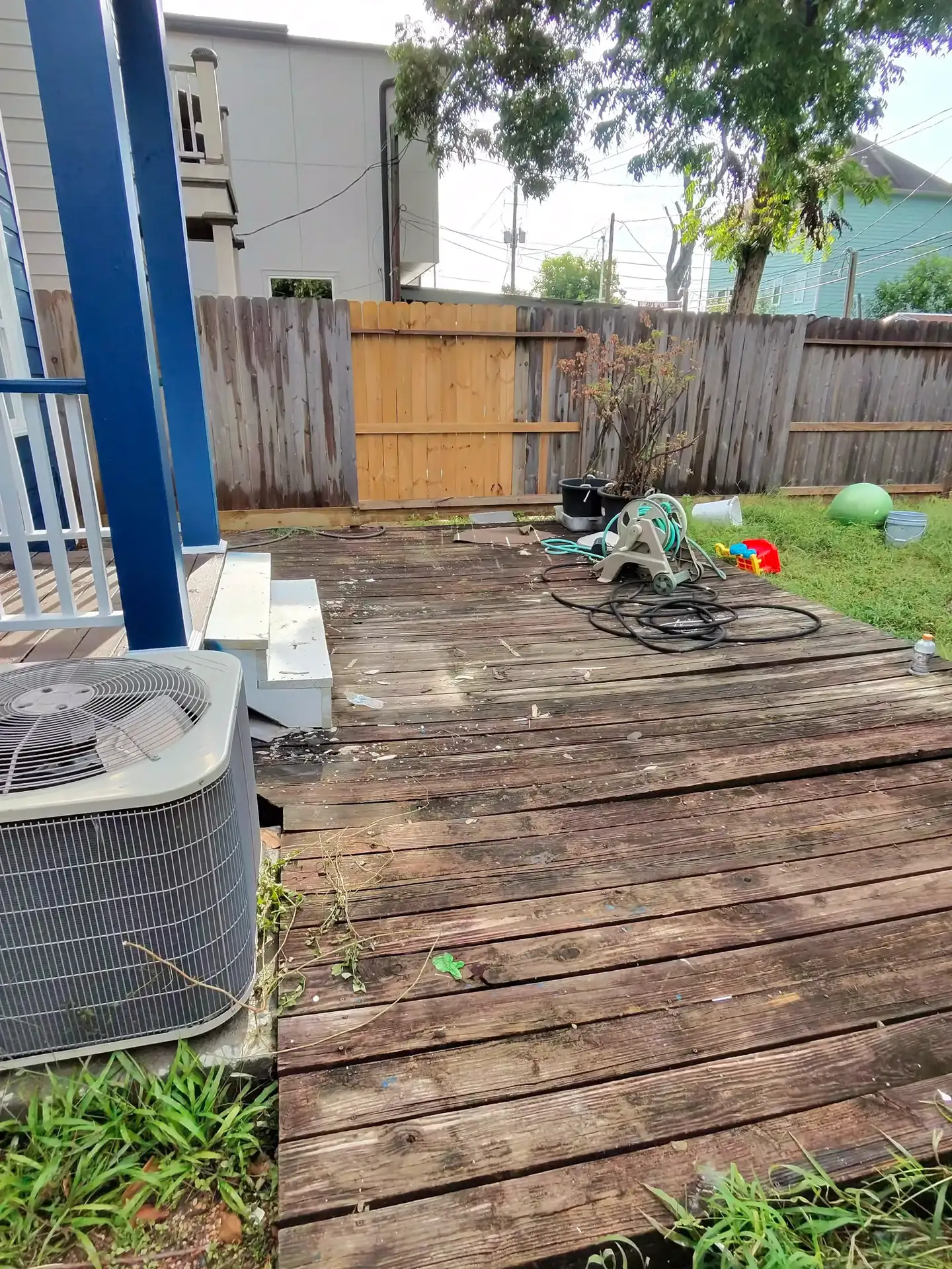 A single new tan board patches a weathered wood fence line along a cluttered back deck in Friendswood, TX.