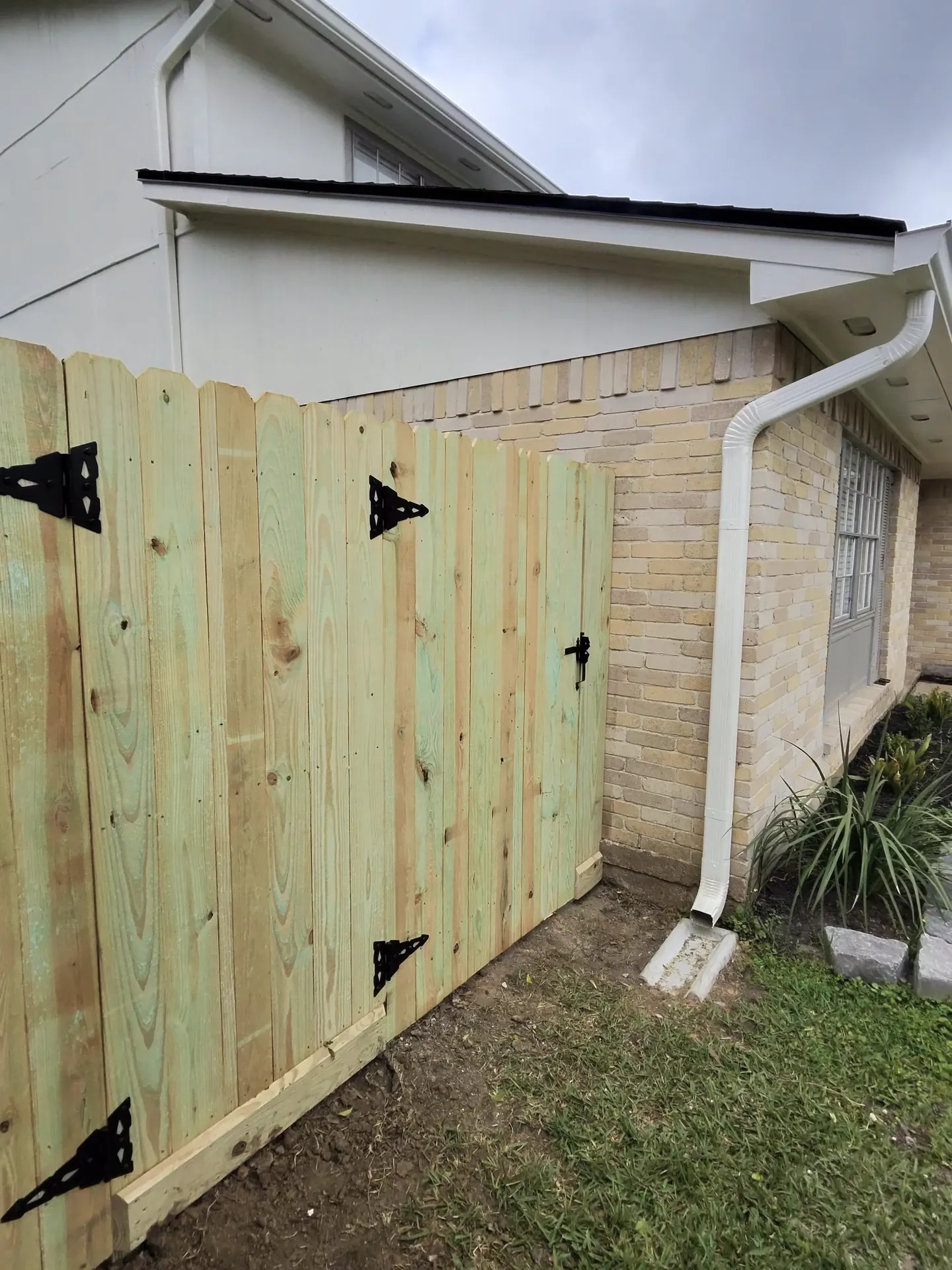 A new pressure-treated wood gate with black decorative strap hinges stands closed beside a brick home in Pearland, TX.