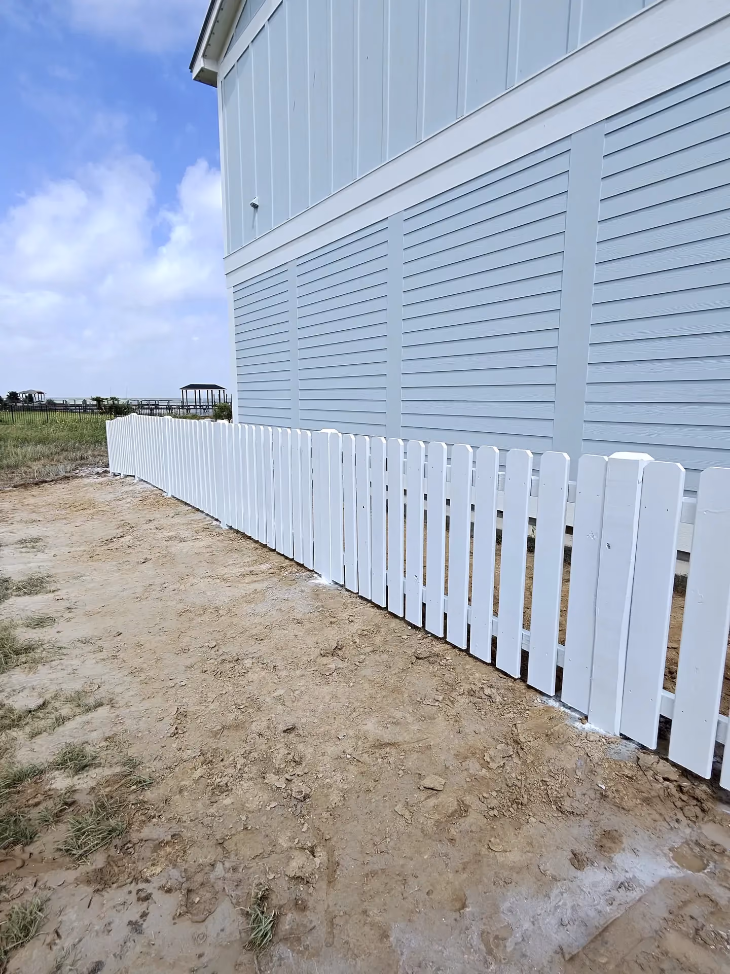 A white picket fence lines the sandy yard of an elevated coastal home with blue-gray siding in Pearland, TX.