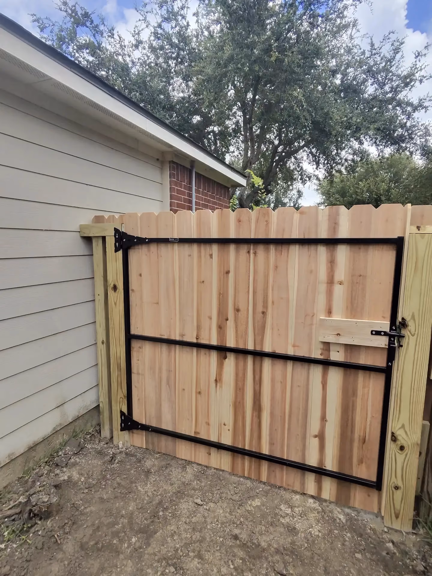 A cedar dog-ear gate with black T-hinges stands beside a beige house on the Texas Gulf Coast.