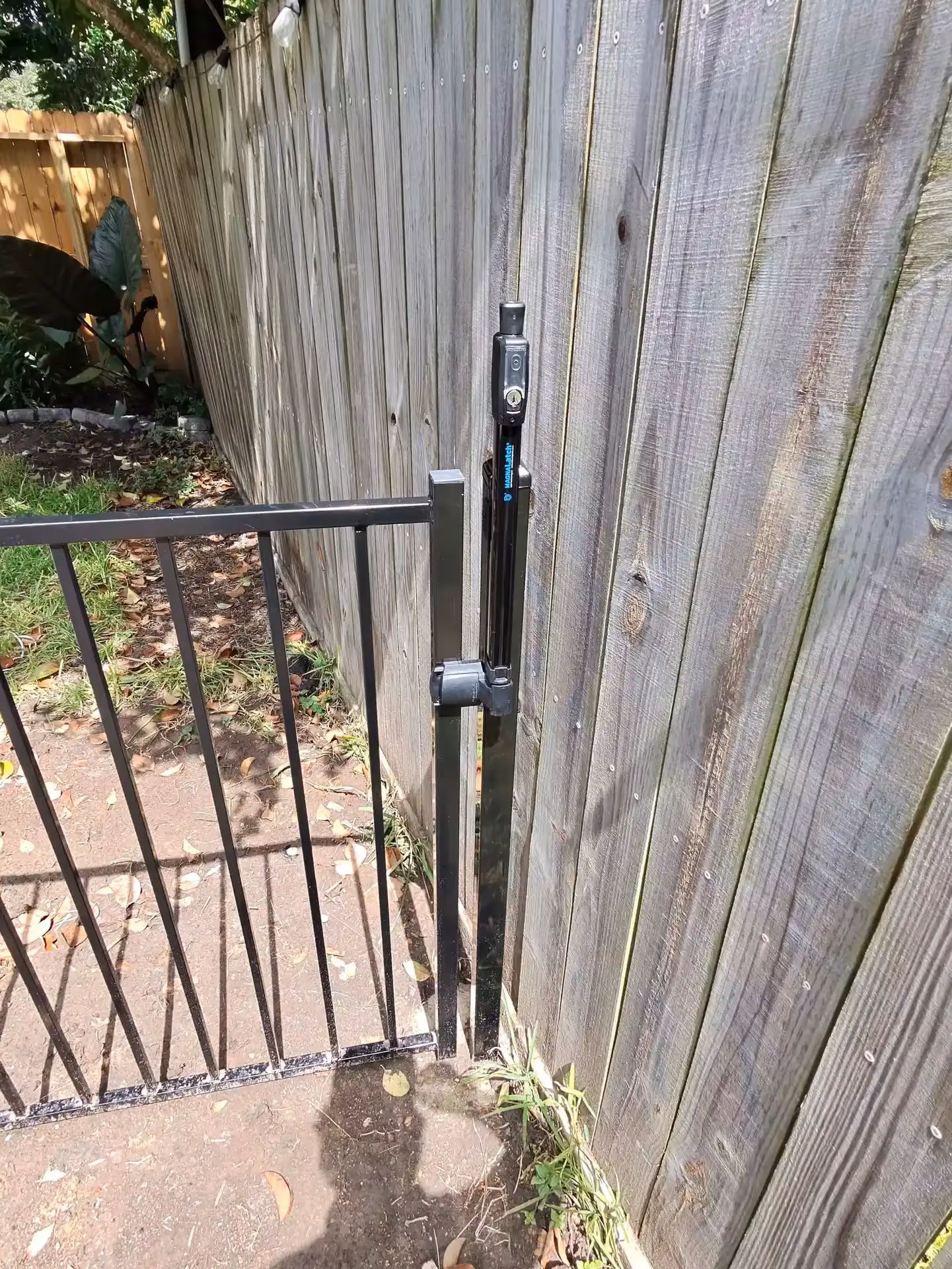 A black aluminum pool gate closes against a weathered wood fence with a MagnaLatch lock on the Texas Gulf Coast.