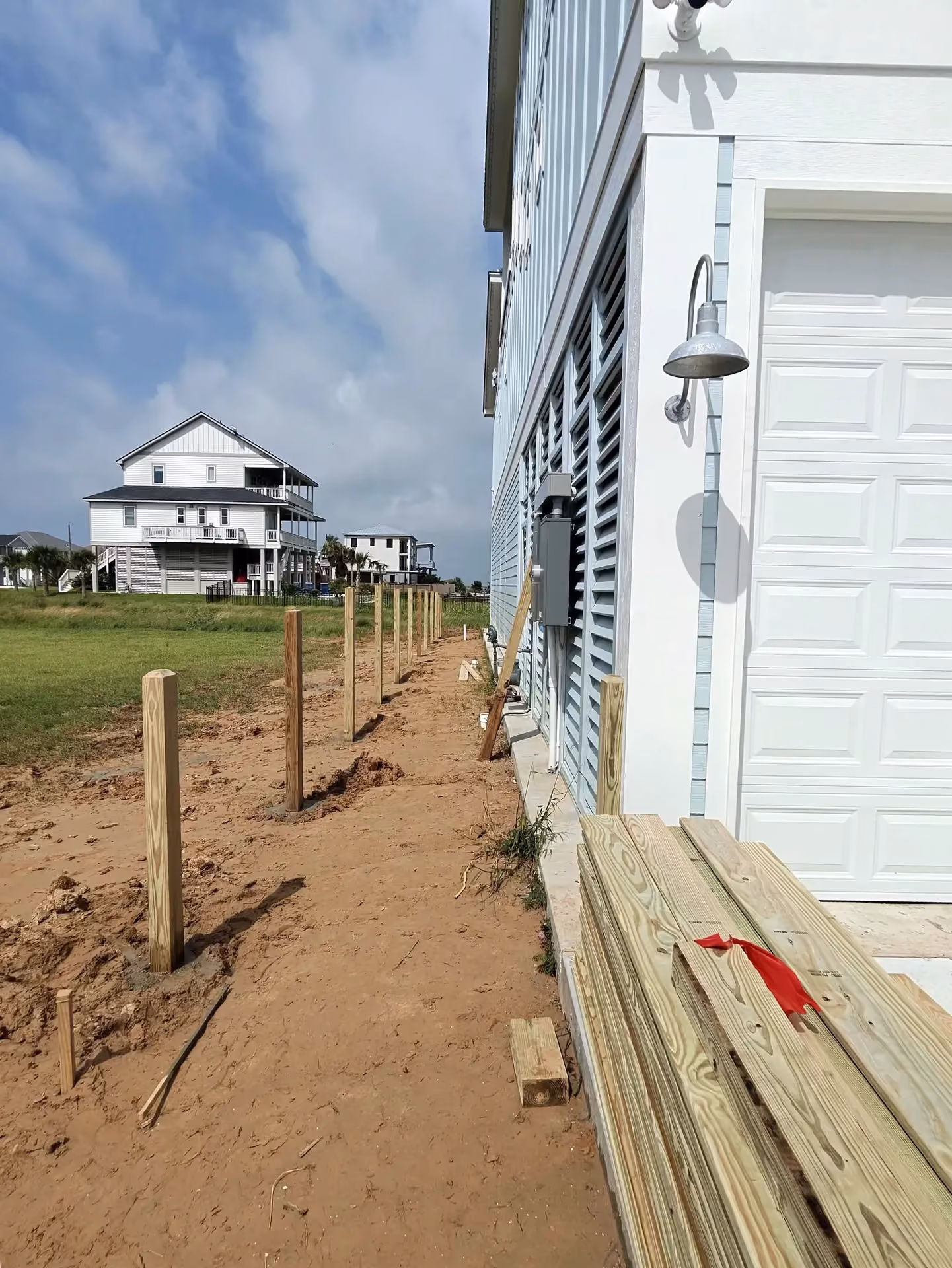 Freshly set pressure-treated fence posts line a sandy yard beside an elevated coastal home on the Texas Gulf Coast.