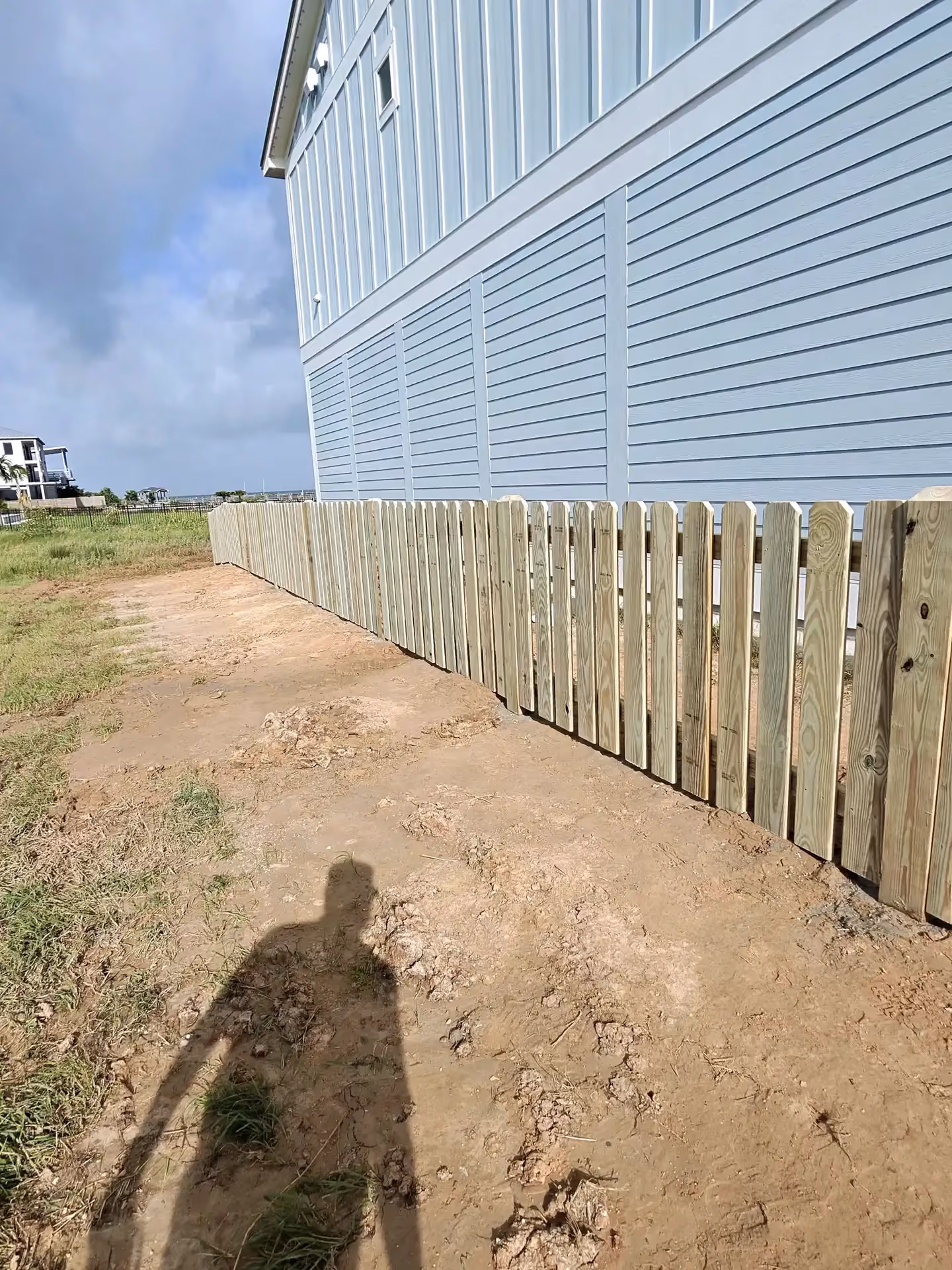 A row of natural wood picket fencing runs beside a blue coastal home on the Texas Gulf Coast.