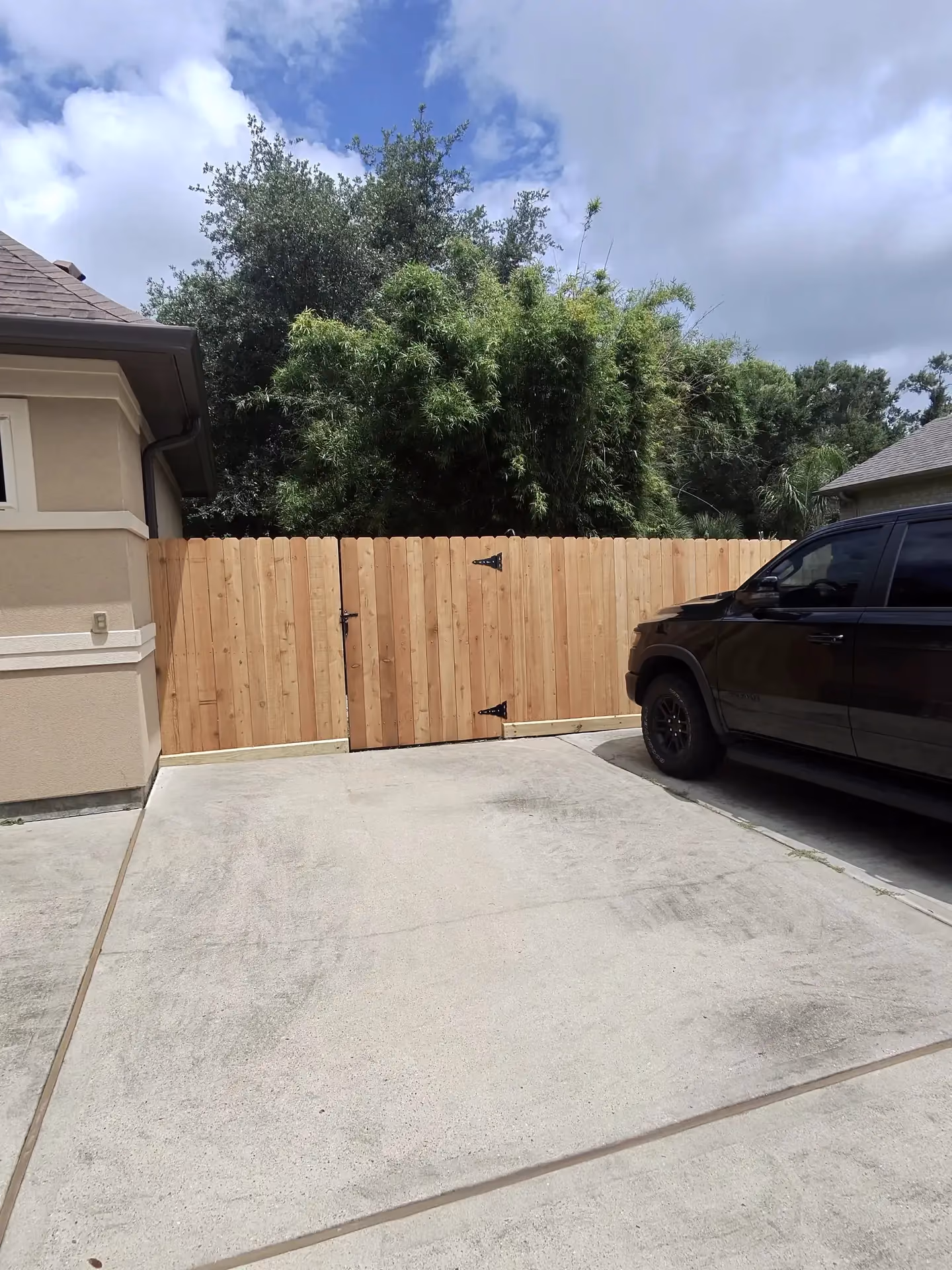 A finished cedar wood gate with black strap hinges secures a driveway beside a stucco home in Houston, TX.