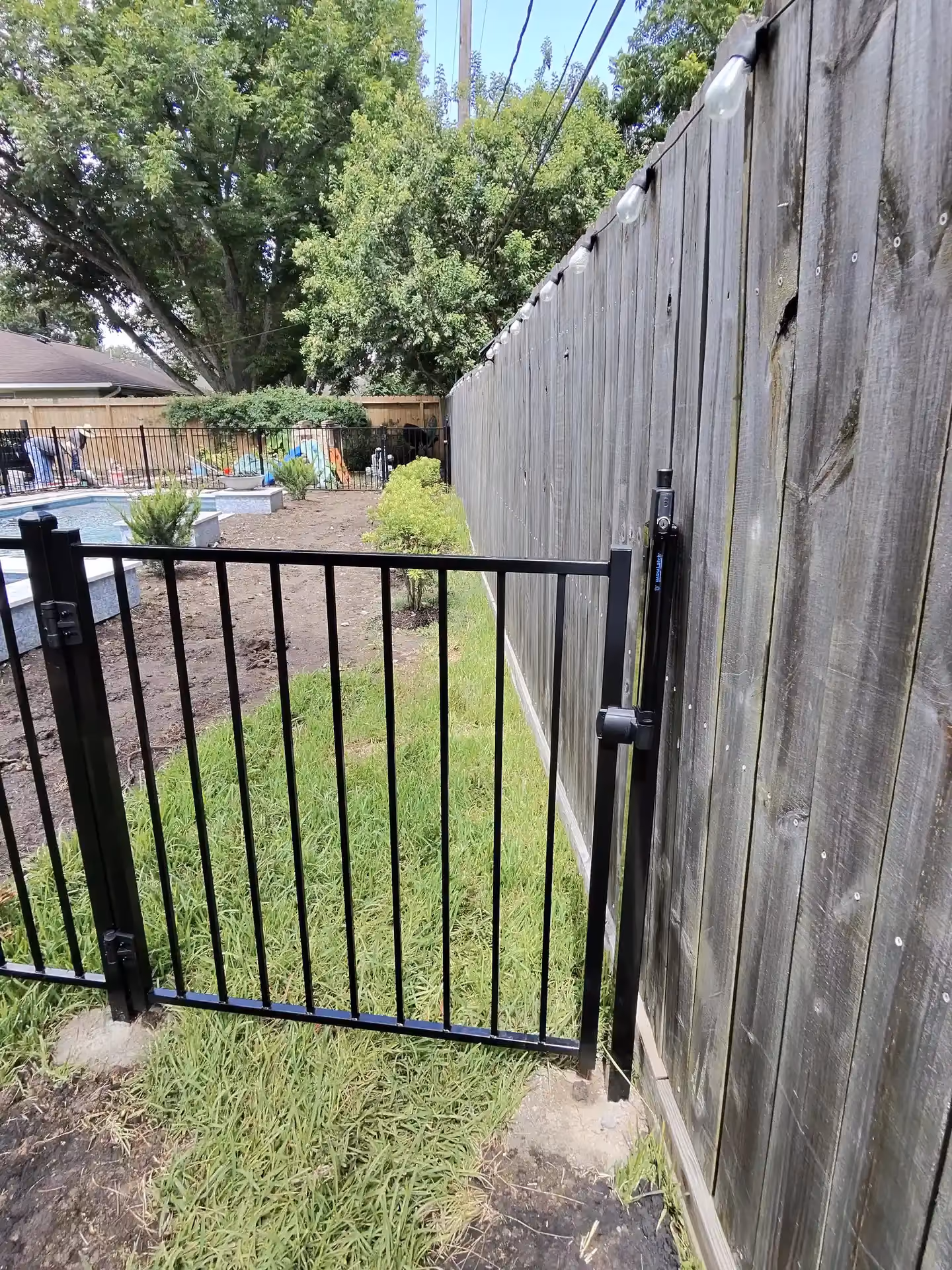 A black aluminum gate opens beside an older wood privacy fence strung with string lights in Houston, TX.