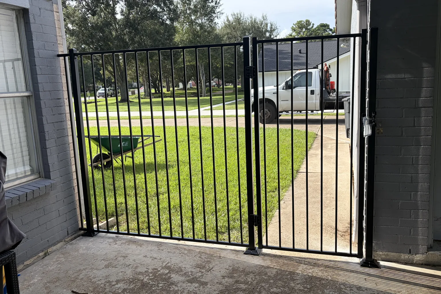 Black metal double gate closing off a home's side patio, with a green front lawn and shade trees beyond
