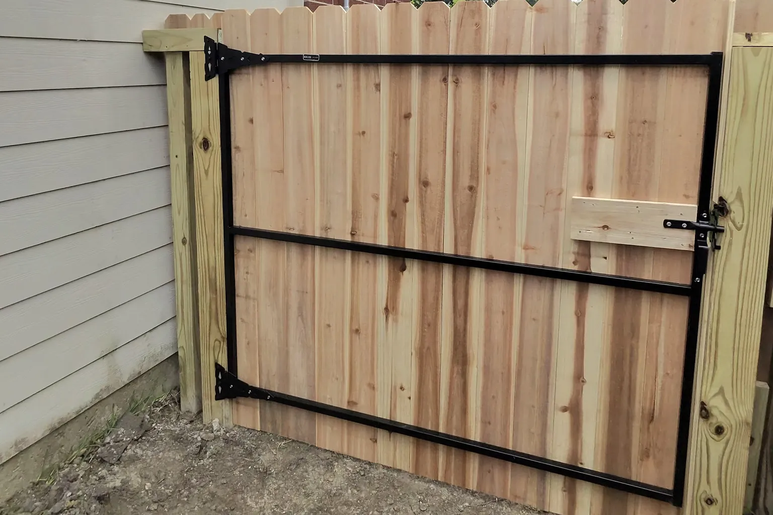 Cedar privacy gate built on a black steel frame, hung from a pressure-treated hinge post beside a home