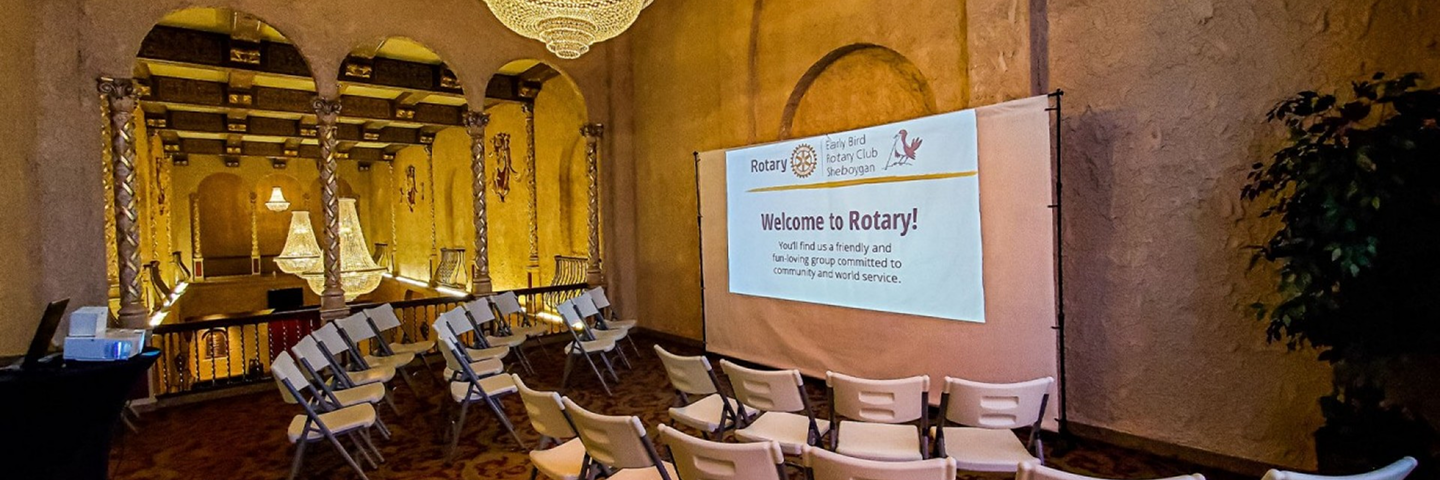 Historic lobby set up with chairs facing a Rotary Club welcome banner and ornate chandeliers.