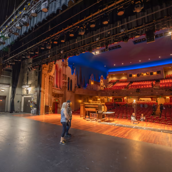 View from stage of empty theater with red seats, ornate balconies, and three people standing on stage.