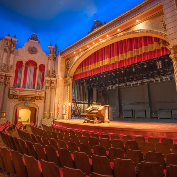 Empty ornate theater with red curtains and grand piano on stage under blue ceiling.