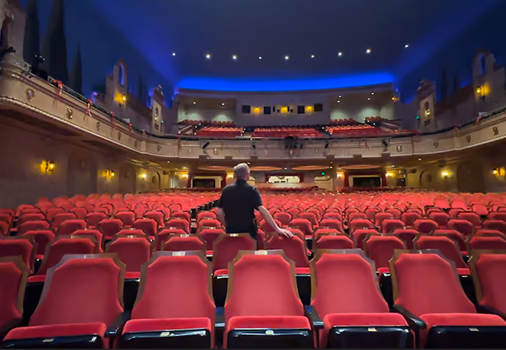 A person sitting alone in the middle of a large empty theater with red seats and blue ceiling lights.