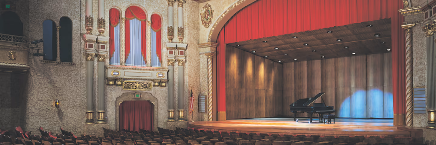 Empty ornate theater with red velvet curtains and a grand piano on stage under spotlights.