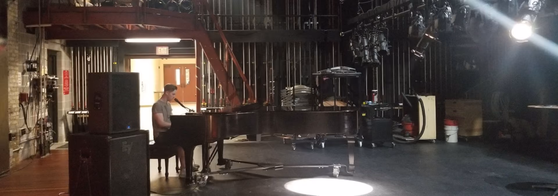 Man playing grand piano on a dimly lit stage with spotlight and equipment around.