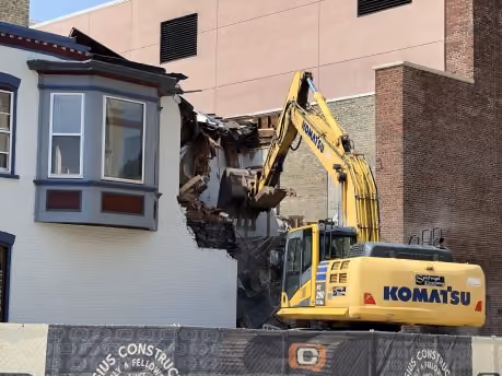 Yellow Komatsu excavator demolishing part of a building with a white and brick facade.