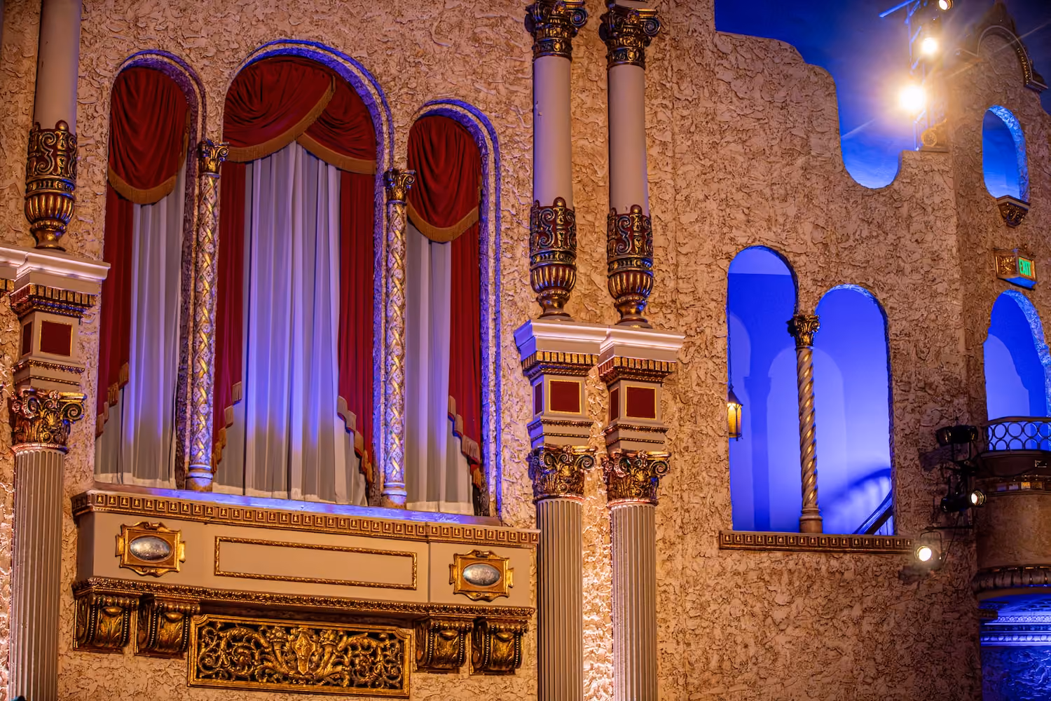 Ornate theater interior with gold accents, red curtains, and blue-lit archways on textured beige walls.