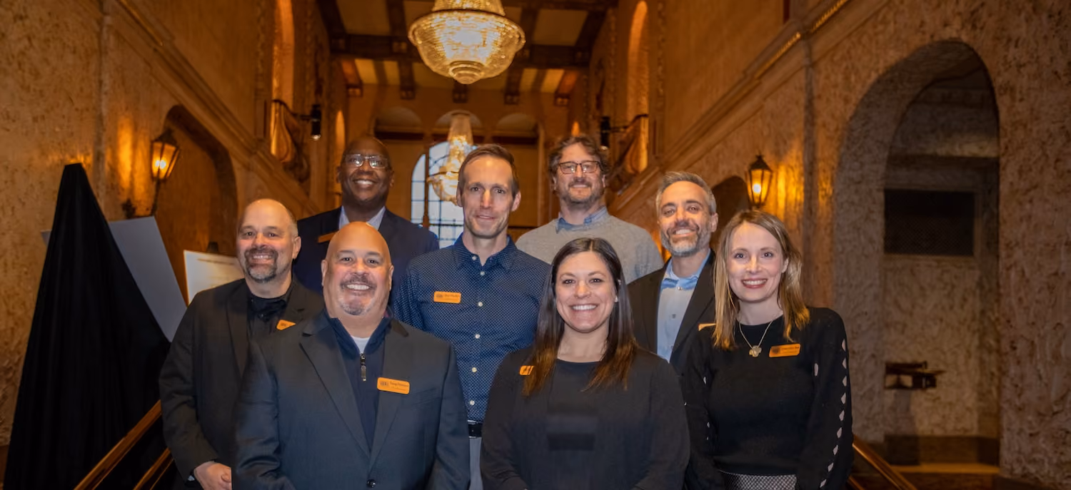 Group of eight people smiling, posed in a warmly lit rustic hallway with chandeliers above.