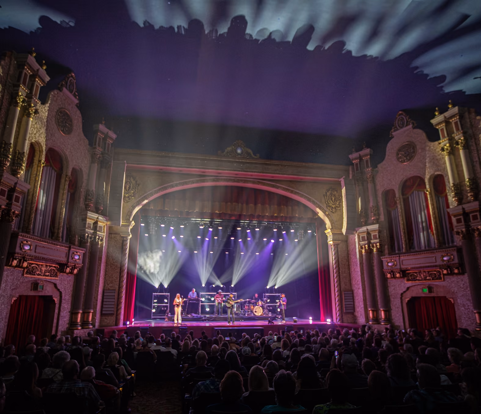 Band performing under purple and white stage lights in a large ornate theater with seated audience.