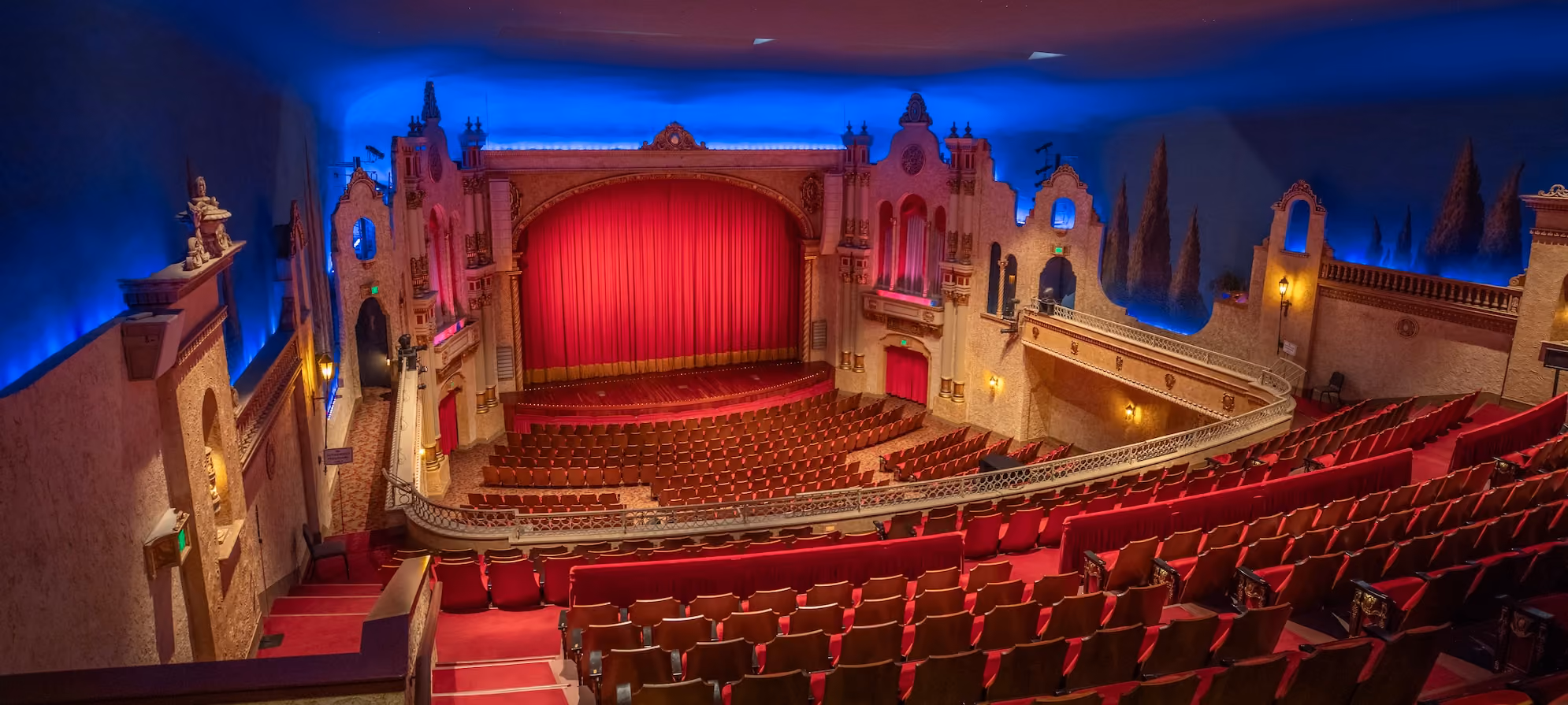 Interior of ornate theater auditorium with red seats and red curtain on stage under blue lighting.