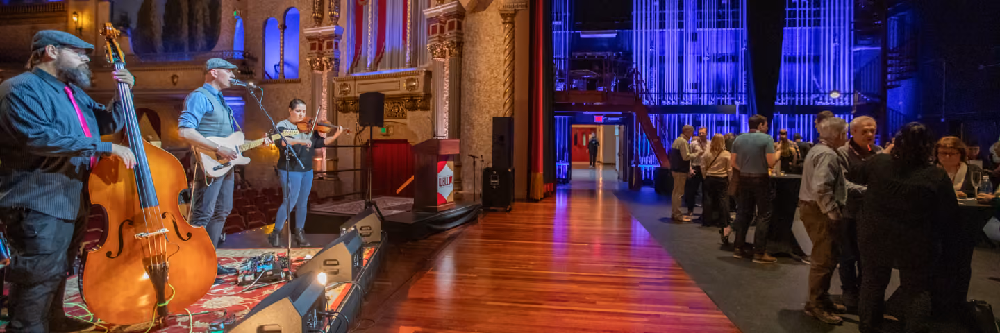 Musicians with upright bass, guitar, and violin perform on stage as people socialize backstage.