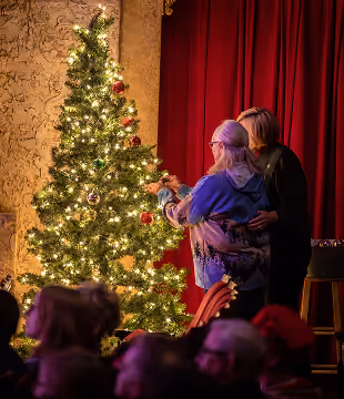 Two women decorate a tall Christmas tree lit with warm white lights on a stage with red curtains.