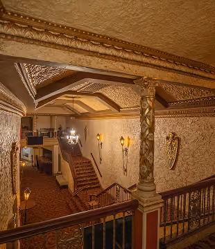 Ornate vintage staircase hall with carved columns, patterned ceiling, and warm wall sconces lighting the space.