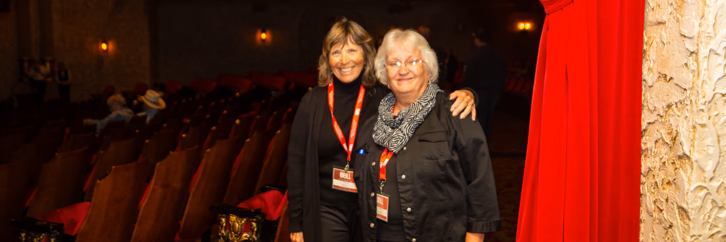 Two smiling women wearing volunteer badges standing with their arms around each other in an auditorium.