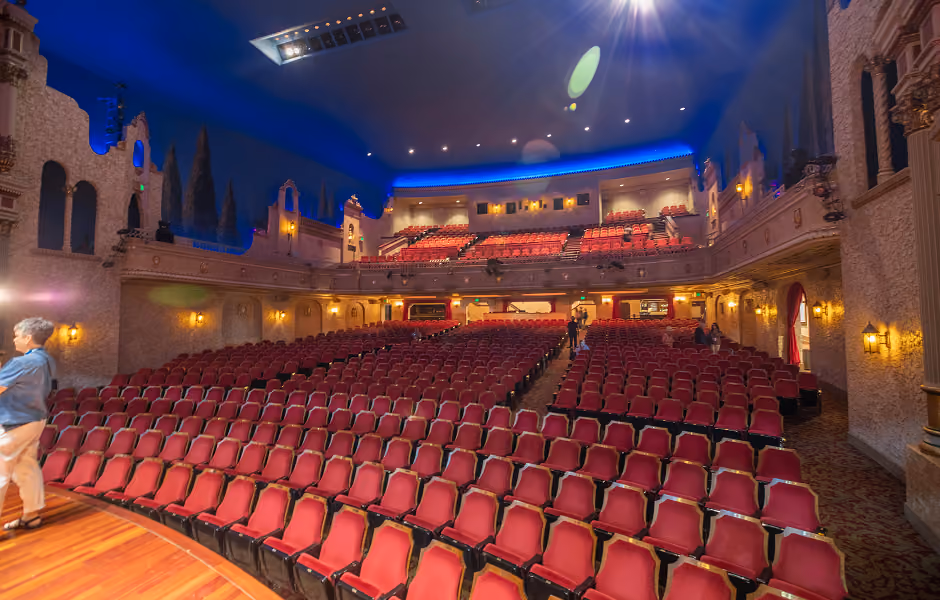 A view from stage-left looking up into the expansive auditorium toward the rear projection booth and upper balcony levels. Lines of red seats sweep across patterned carpeting, flanked by warm wall sconces and Mediterranean architectural elements.