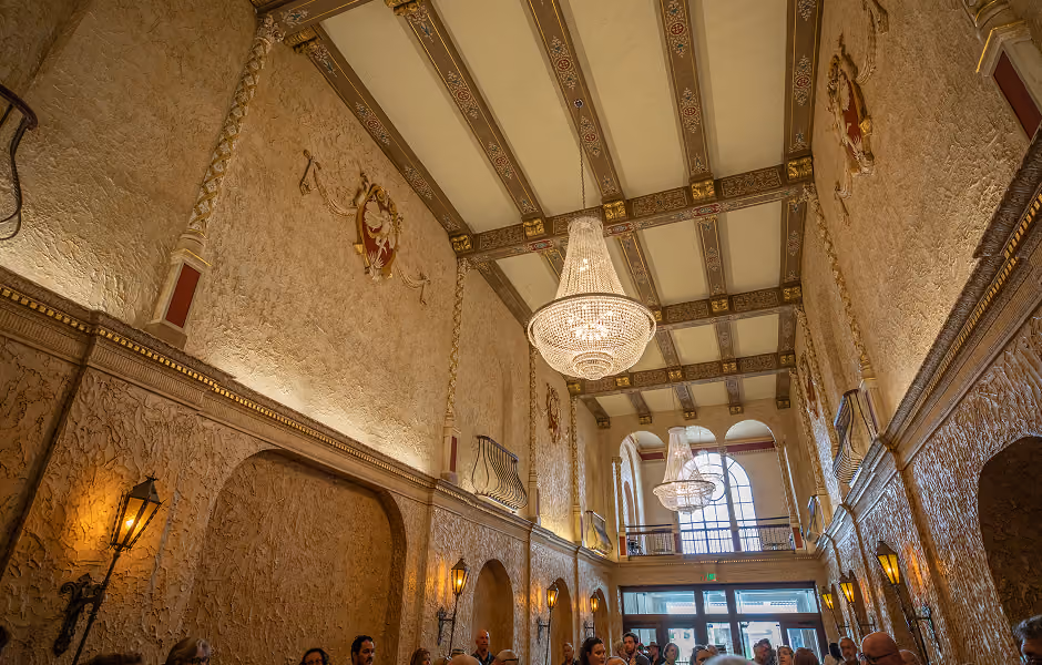 A wide, low-angle view looking up toward the high, ornate ceiling of a grand lobby. Large crystal chandeliers illuminate textured stucco walls decorated with golden crests, faux balconies, decorative wall sconces, and double doors at the far end under a large arched window.
