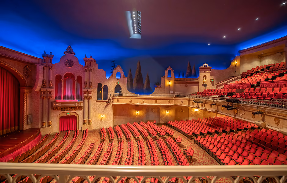 A high-angle view over a white balcony rail capturing the main floor seating arrangement, side organ chamber facade, and upper balcony section under a deep blue ceiling with atmospheric lighting.