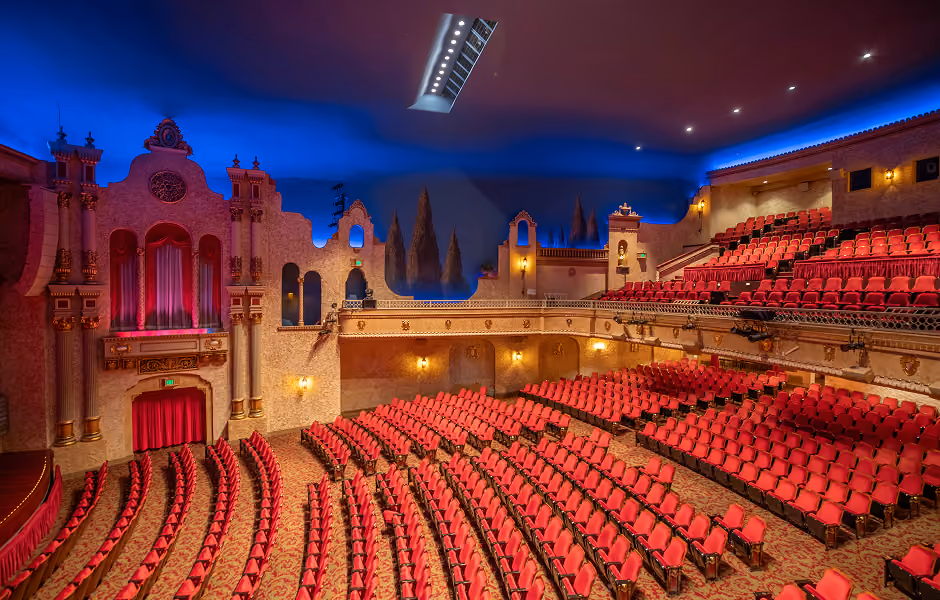 A high-angle interior view of an atmospheric theater auditorium showing curved rows of red seats, side wall facades with painted cypress trees, and an upper balcony tier under a dark blue painted sky ceiling.