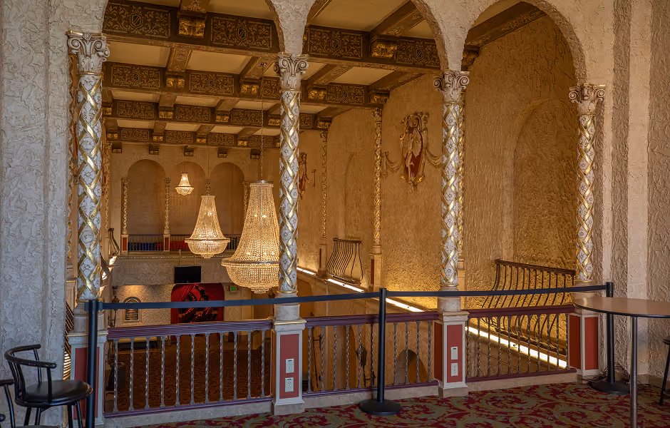 An upper-balcony view of a grand interior space framed by three arched openings supported by white and gold detailed columns. Crystal chandeliers hang in the open void beyond a dark railing, overlooking lower-level arched doorways and textured walls.