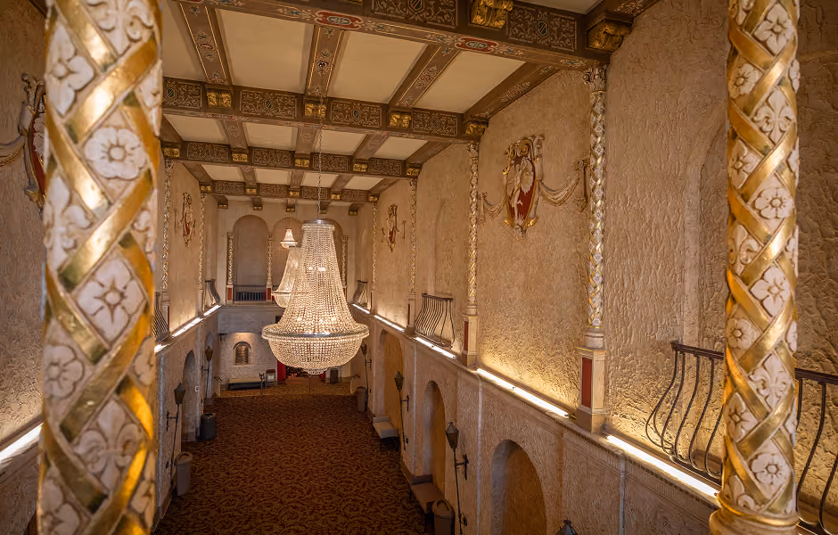 A high-angle perspective from between two elaborate gold and white spiral columns, looking down the length of an ornate lobby hallway. Large crystal chandeliers hang centrally above a long red patterned carpet flanked by arched doorways, lantern wall sconces, and decorative wall crests.