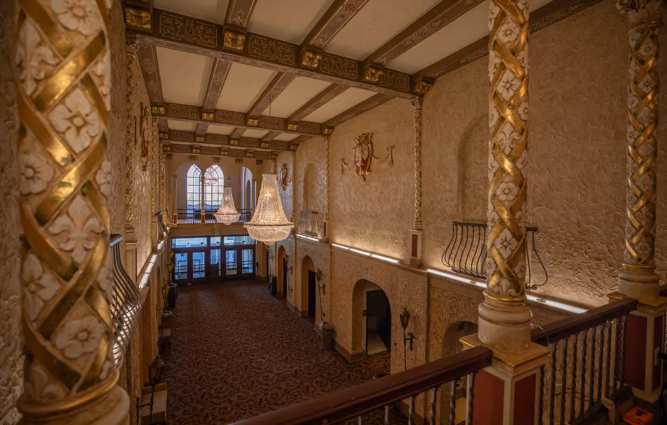 An elevated, high-angle view looking down into a long grand lobby, framing ornate gold spiral-patterned columns in the foreground. Sparkling crystal chandeliers hang beneath a detailed coffered ceiling, looking toward glass entrance doors at the end of a patterned red carpet.