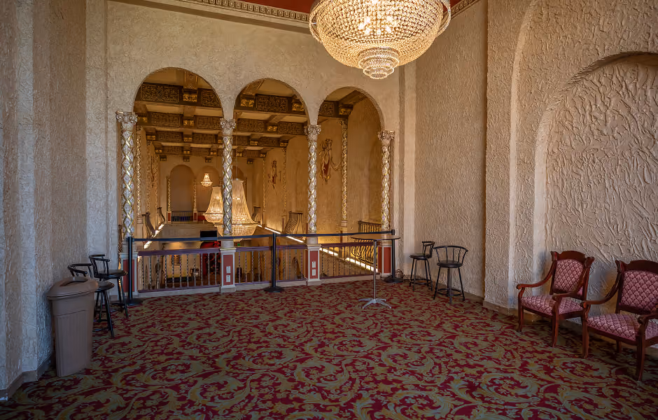 An upper-level seating or mezzanine area with red and gold patterned carpeting, featuring a large crystal chandelier hanging overhead. Detailed golden columns separate the space from a lower lobby view, with two pink upholstered armchairs placed along a textured stucco wall on the right.