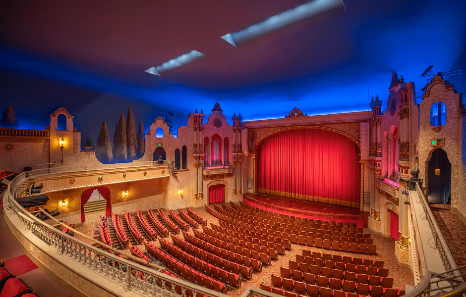 An elevated balcony perspective overlooking the entire auditorium, showing curved rows of plush red seats leading to a stage with a closed red main curtain. Ornate side arches, cypress tree murals, and a painted blue ceiling surround the seating area.