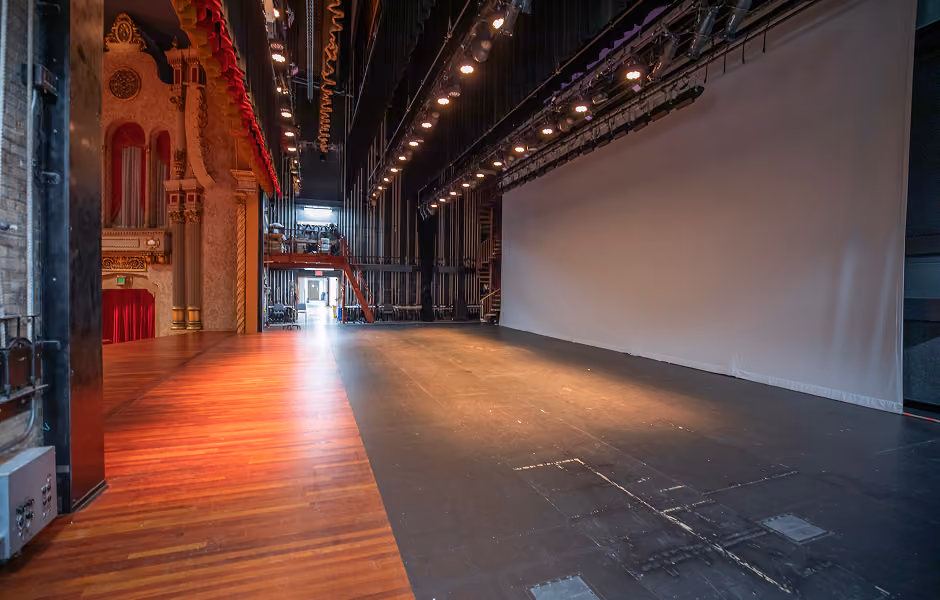 A side-angle perspective of the backstage area and dark stage floor, featuring a large white backdrop projection screen hanging opposite the polished wooden apron leading to the main theater hall.